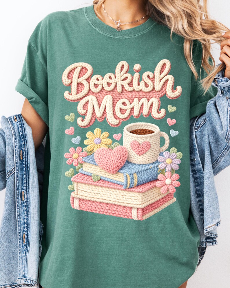 May include: Green t-shirt with the text "Bookish Mom" in a knitted style. The design includes a stack of books, a coffee mug, flowers, and hearts. The shirt has rolled-up sleeves and is worn with a denim jacket.