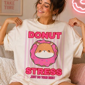 Könnte beinhalten: Cremefarbenes T-Shirt mit einem rosa und braunen Donut-Motiv. Die Grafik zeigt einen Cartoon-Hamster in einem rosa glasierten Donut mit Streuseln. Der Text "DONUT STRESS" und "JUST DO YOUR BEST" ist in Rosa gedruckt.