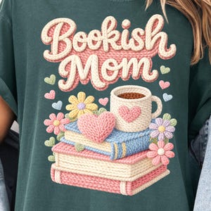 May include: Dark teal t-shirt with the words "Bookish Mom" in a 3D knit design. The graphic includes a stack of books, a coffee mug, hearts, and flowers, all in a soft, pastel color palette.