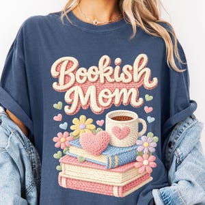 May include: Navy blue t-shirt with the text "Bookish Mom" in a knitted design. The graphic includes a stack of books, a coffee mug with a heart, flowers, and hearts.
