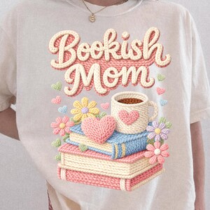May include: Cream-colored t-shirt with the text "Bookish Mom" in a 3D knit design. The graphic includes a stack of books, a coffee mug, flowers, and hearts. The design is in pastel colors.