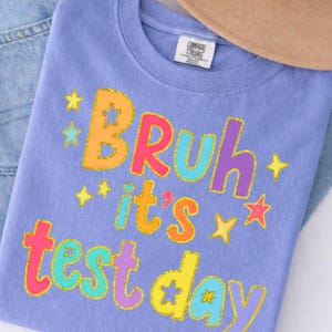 Op de afbeelding: Een lila t-shirt met de tekst "Bruh it's test day" in kleurrijke, met glitter omlijnde letters en sterren. Het shirt is opgevouwen op een spijkerbroek. Het ontwerp is speels.