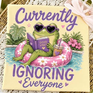 Könnte beinhalten: Leinwanddruck mit einer Cartoon-Froschfigur, die eine herzförmige Sonnenbrille trägt und in einem Pool auf einem rosa Schwimmring schwimmt. Der Frosch liest ein Buch mit "Good Vibes". Der Text "Currently Ignoring Everyone" ist auf der Leinwand geschrieben.