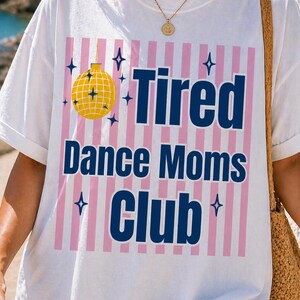 Könnte beinhalten: Weißes T-Shirt mit dem Text "Tired Dance Moms Club" in Marineblau. Das Design umfasst eine gelbe Discokugel und rosa vertikale Streifen. Blaue Sternakzente sind ebenfalls vorhanden.
