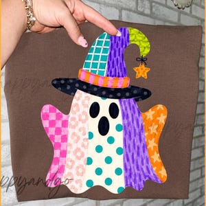 Könnte beinhalten: Ein braunes T-Shirt mit einem farbenfrohen Geisterdesign. Der Geist trägt einen Patchwork-Hexenhut mit einem Sternanhänger. Der Körper des Geistes ist ein Patchwork aus rosa, weißen, türkisfarbenen, lila und orangefarbenen Mustern. Das Design ist skurril und festlich.