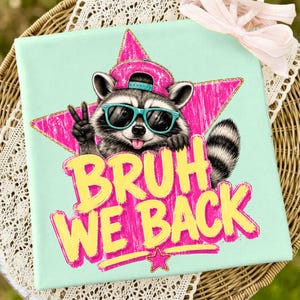 Puede incluir: Un lienzo cuadrado de color verde menta con el gráfico de un mapache que lleva una gorra rosa y gafas de sol turquesas, haciendo el signo de la paz. El texto BRUH WE BACK está impreso en amarillo con borde rosa sobre una estrella rosa.
