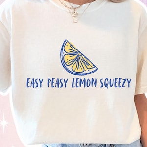 Puede incluir: Camiseta color crema con un gráfico de rodaja de limón amarillo y azul y el texto azul "EASY PEASY LEMON SQUEEZY". Un estilo casual y cómodo.