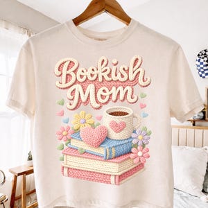 May include: Cream-colored t-shirt with the text "Bookish Mom" in a 3D design. The graphic features a stack of books, a coffee mug with a heart, flowers, and hearts. The design is in shades of pink, blue, yellow, and green.