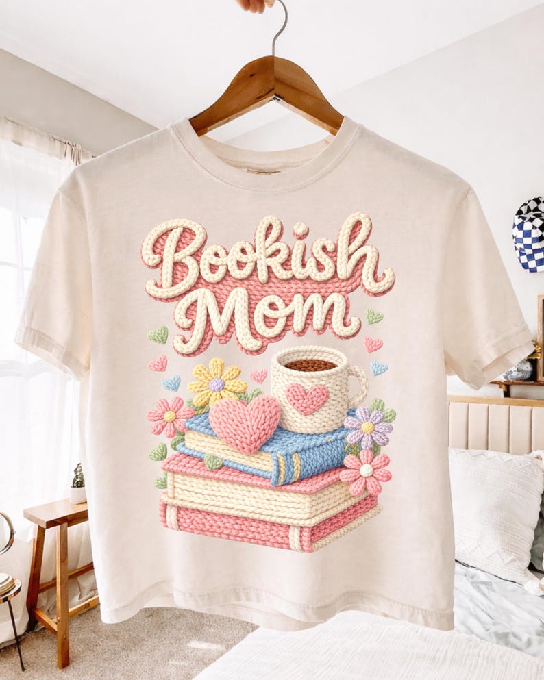 May include: Cream-colored t-shirt with the text "Bookish Mom" in a 3D design. The graphic features a stack of books, a coffee mug with a heart, flowers, and hearts. The design is in shades of pink, blue, yellow, and green.