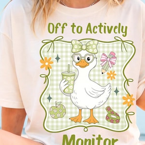 May include: Cream-colored t-shirt with a cartoon duck wearing glasses and a bow, holding a drink. The shirt features the text "Off to Actively Monitor" in green, along with other graphics like a pencil, apple, and flowers. The design is framed by a green border.