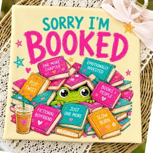 Könnte beinhalten: Eine quadratische Leinwand mit dem Text "SORRY I'M BOOKED" in Türkis und Pink. Das Bild zeigt einen Frosch, der aus einem Stapel bunter Bücher hervorlugt. Ein Getränk mit Strohhalm befindet sich in der linken unteren Ecke. Das Thema ist Lesen und Bücher.
