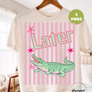 Könnte beinhalten: Cremefarbenes T-Shirt mit einer Cartoon-Alligator-Illustration. Das Design beinhaltet das Wort "Later" in Grün und Rosa, rosa Sterne und rosa vertikale Streifen. Das Shirt hängt an einem Holzbügel.