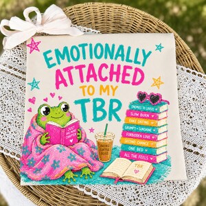 May include: A square canvas print with the text "EMOTIONALLY ATTACHED TO MY TBR" in teal and pink. A cartoon frog reads a book, wrapped in a pink blanket. A stack of books and a coffee cup are also shown. The print is on a wicker basket.