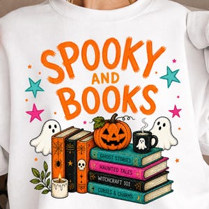 Könnte beinhalten: Ein weißes Sweatshirt mit dem orangefarbenen Text "SPOOKY AND BOOKS" über einem Stapel bunter Bücher, einem Kürbis, einer dampfenden Tasse mit Geister-Symbol, zwei weißen Geistern, einer brennenden Kerze sowie kleinen pinken und türkisen Sternen.
