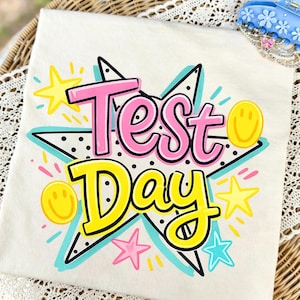 Könnte beinhalten: Cremefarbenes T-Shirt mit einem farbenfrohen Aufdruck "Test Day". Das Design zeigt den Schriftzug "Test" in Pink und "Day" in Gelb, umrandet von Schwarz, mit Sternen und Smiley-Gesichtern.