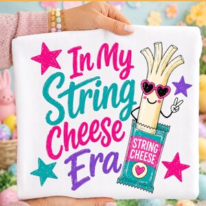 Könnte beinhalten: Ein weißes T-Shirt mit einer Grafik eines Cartoon-Käsesticks, der herzförmige Sonnenbrillen trägt und ein Friedenszeichen macht. Der Text In My String Cheese Era ist in Pink, Türkis und Lila geschrieben, umgeben von farbigen Sternen.