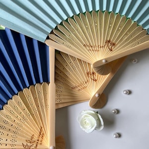 Può includere: Diversi ventagli di carta aperti in tonalità blu e beige. I ventagli beige hanno un manico in legno e sono incisi con nomi e date. Una rosa bianca e perle sparse completano la composizione.