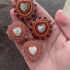 May include: Three beaded heart-shaped ornaments with a white shell center. The ornaments are surrounded by red, pink, and gold beads.