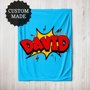 Puede incluir: Una manta azul claro hecha a medida con el nombre "DAVID" en letras rojas grandes al estilo cómic. El nombre está sobre un estallido amarillo con contornos y acentos negros. La manta tiene un fondo azul claro liso.