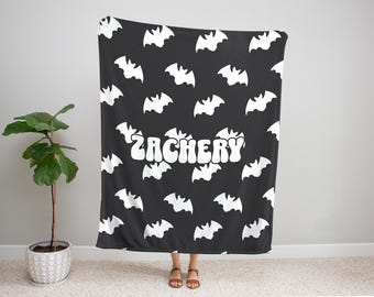 Può includere: Coperta nera con un motivo di pipistrelli bianchi e il nome "ZACHERY" in bianco, con un carattere retrò. La coperta è tenuta in alto, con il bordo inferiore vicino al pavimento. I pipistrelli sono distribuiti uniformemente.