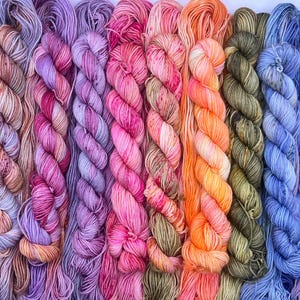 May include: A collection of ten hand-dyed yarn skeins arranged side-by-side in a vertical row. The skeins feature a variegated colour palette including shades of deep red, soft pink, vibrant magenta, lavender, bright orange, olive green, and blue.