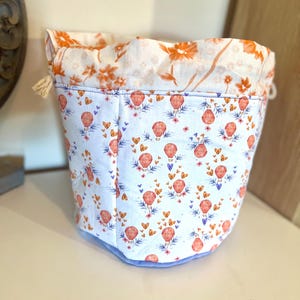 May include: A fabric storage basket with a white base, patterned with orange hot air balloons, butterflies, and flowers. The top edge has an orange and white floral print. The basket has a light blue base and a drawstring closure.