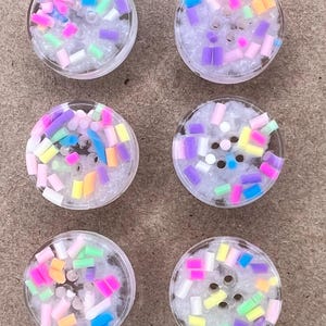 May include: Six circular, clear buttons featuring a textured, white, glittery base embedded with an assortment of small, cylindrical, multi-coloured sprinkles in shades of pink, blue, yellow, purple, and green. Each button has four holes.