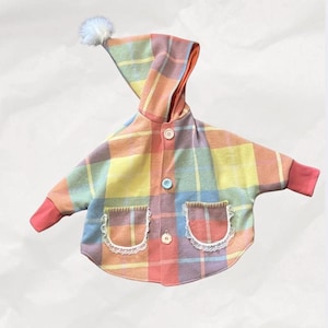 May include: A hooded jacket featuring a multi-colored plaid pattern in shades of yellow, orange, blue, and purple. The garment includes three front buttons, two pockets with white lace trim, solid coral-colored cuffs, and a white pom-pom on the hood.