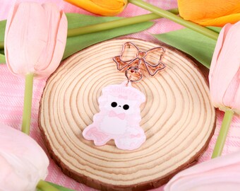 May include: A pink and white resin charm of a cartoon animal wearing a bow tie and a crown. The charm is attached to a rose gold metal key chain with a bow.