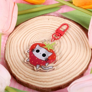 Può includere: Un portachiavi a forma di gatto rosso e grigio con un design a fragola. Il portachiavi è in acrilico trasparente e ha un moschettone in metallo rosso.