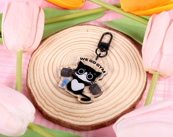 May include: Black and white cat keychain with a dumbbell in each paw. The keychain is made of clear acrylic and has the text "WE GO GYM" printed on it. The keychain is on a wooden surface with pink tulips in the background.