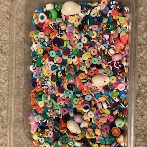 May include: A clear plastic container filled with a variety of colourful beads. The beads are made of plastic and come in a variety of shapes and sizes. Some of the beads are round, some are square, and some are shaped like letters. The beads are all different colours, including red, blue, green, yellow, orange, purple, and pink.