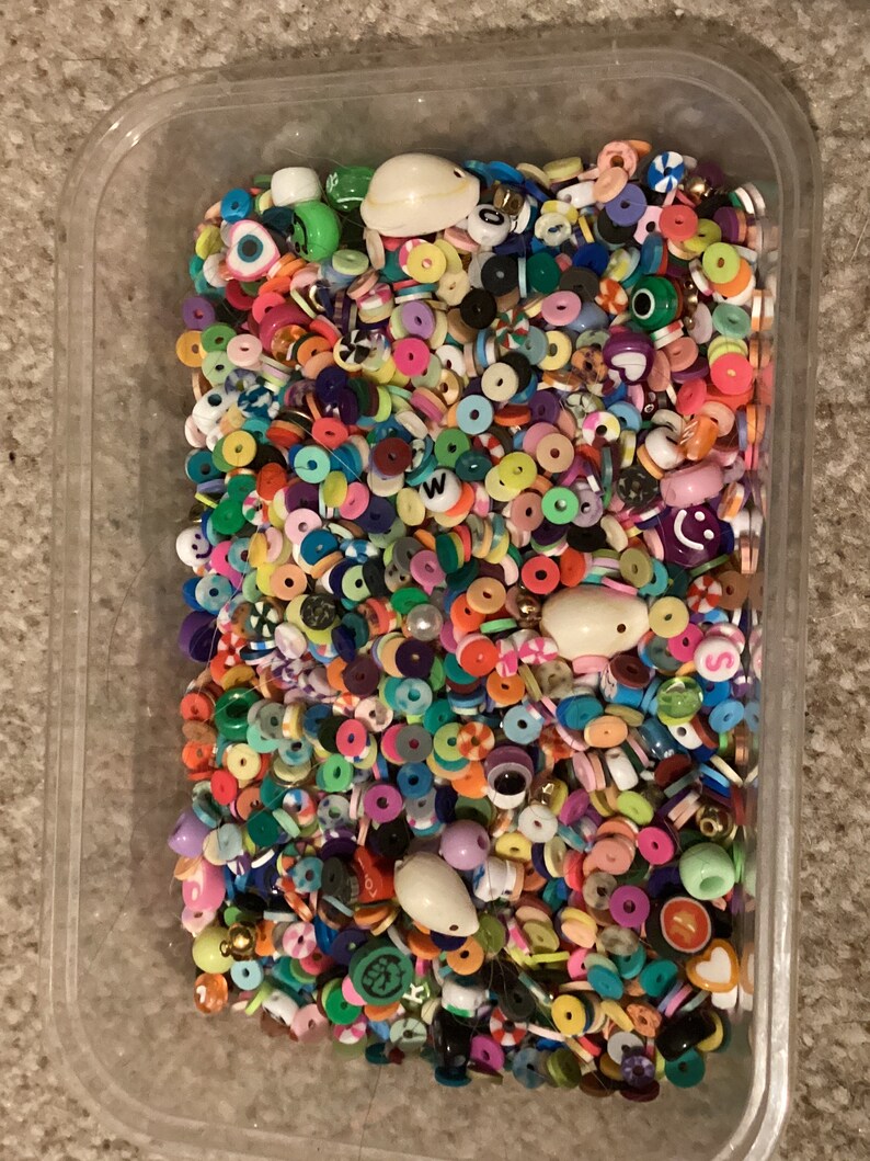 May include: A clear plastic container filled with a variety of colorful beads. The beads are made of plastic and come in a variety of shapes and sizes. Some of the beads are round, some are square, and some are shaped like letters. The beads are all different colors, including red, blue, green, yellow, orange, purple, and pink.