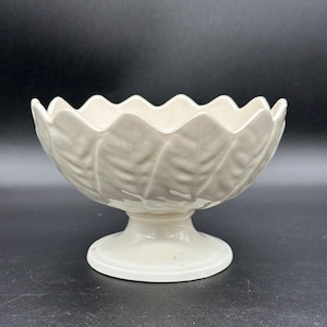 Puede incluir: Cuenco de cerámica blanco roto con pie y borde festoneado. El cuenco presenta un diseño de hojas y se apoya sobre una base corta. El cuenco tiene unos 15 cm de diámetro y 10 cm de altura.