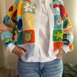 Peut inclure: Un cardigan en crochet blanc avec des carrés colorés présentant différents motifs, notamment une fleur, une grenouille, un champignon, un ours et une étoile de mer.