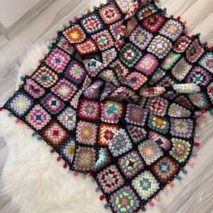 Puede incluir: Una colorida manta de ganchillo hecha a mano con un diseño de retazos de motivos cuadrados en varios colores. La manta tiene un borde negro y está adornada con pequeñas borlas de colores en los bordes. La manta está sobre una superficie blanca y esponjosa.