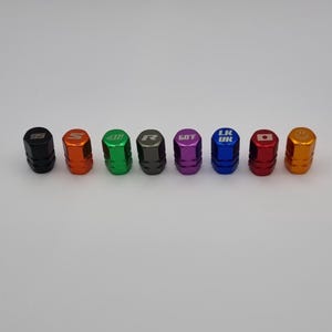 May include: Eight different colored tire valve stem caps with various designs and logos. The caps are black, orange, green, gray, purple, blue, red, and gold.