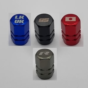 May include: A collection of five metal tire valve stem caps in various colors. The caps are blue, black, red, and gray. The blue cap has "LK UK" in white. The black cap has "95" in silver. The red cap has a Japanese flag. The gray cap has an "R".