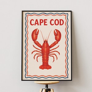Op de afbeelding: Een ingelijst print met een illustratie van een rode kreeft en de tekst "CAPE COD" in rode letters op een witte achtergrond met blauwe en witte golvende randen.