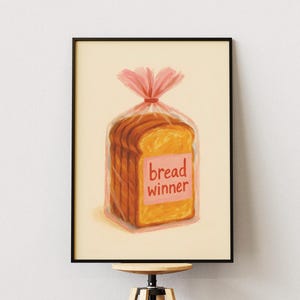 May include: A framed art print depicting a loaf of sliced bread in a clear plastic bag, tied with a pink ribbon. The bread has a pink label that reads "bread winner". The artwork is displayed on a wooden stool.