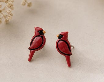 May include: A pair of stud earrings shaped like red cardinals. Each bird features a glossy red body, a black face mask around the eye, and a small yellow beak. The earrings are designed with a post back for attachment, showcasing a detailed bird silhouette.