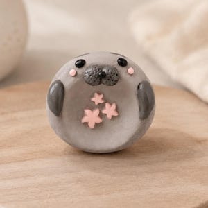 May include: A round, gray clay figurine shaped like a seal with a textured dark gray snout, small black eyes, and pink circular cheeks. The seal has two dark gray flippers on its sides and three small, light pink star-shaped accents on its belly.