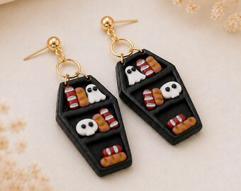 May include: A pair of dangle earrings featuring black coffin-shaped charms. Each charm contains miniature shelves holding tiny white ghosts, white skulls, and small brown and red loaves of bread. The charms hang from gold-toned metal posts.