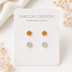 May include: Two pairs of stud earrings on a white display card labeled EARLOOM CREATIONS handcrafted jewelry. The top pair features mustard yellow sun shapes, while the bottom pair consists of light gray circular studs with a white star design.