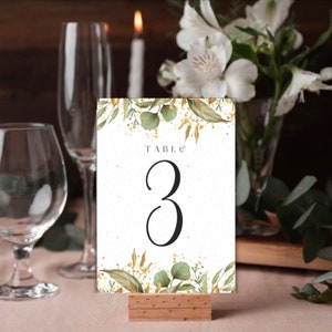 Puede incluir: Una tarjeta de número de mesa blanca con follaje dorado y verde y purpurina dorada. La tarjeta dice "TABLE 3" en texto negro.