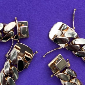 May include: Close-up of a gold chain bracelet with a clasp. The chain is made of thick, interlocking links. The bracelet is lying on a purple surface.