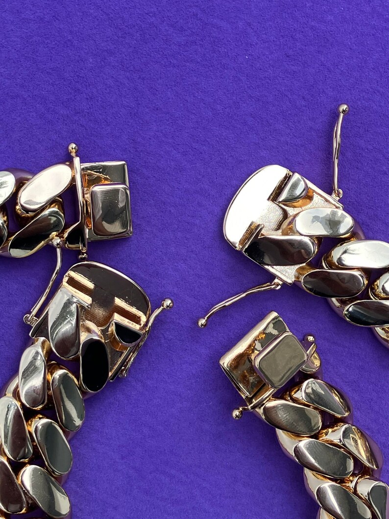 May include: Close-up of a gold chain bracelet with a clasp. The chain is made of thick, interlocking links. The bracelet is lying on a purple surface.