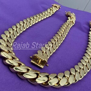 May include: Two gold-coloured curb chains, one longer than the other, are laid out on a purple surface. The chains are made of thick, interlocking links.