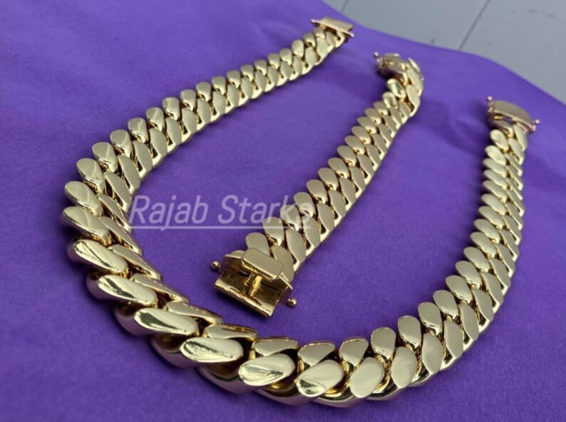 May include: Two gold-coloured curb chains, one longer than the other, are laid out on a purple surface. The chains are made of thick, interlocking links.