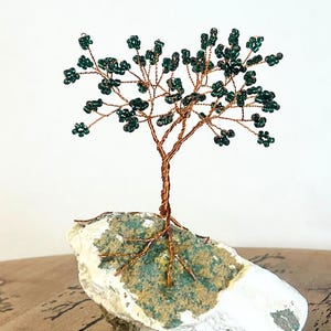May include: A handcrafted wire tree sculpture with a copper-colored trunk and branches, adorned with dark green beads as leaves. The tree is mounted on a white and green stone base, creating a unique decorative piece.